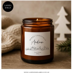Puede incluir: Un tarro de vela de vidrio marrón con una etiqueta blanca que dice "Merry Christmas Andrew" con una ilustración de un pueblo. La vela tiene una mecha encendida y está junto a una decoración de árbol de Navidad de madera blanca.