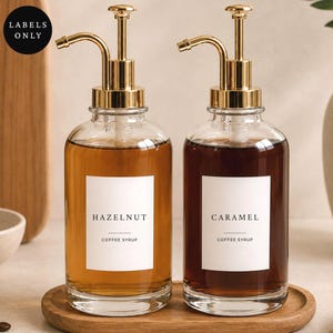 Op de afbeelding: Twee doorzichtige glazen flessen met goudkleurige metalen pompen. Elke fles heeft een wit rechthoekig etiket met zwarte tekst. Eén etiket vermeldt HAZELNUT COFFEE SYRUP en het andere etiket vermeldt CARAMEL COFFEE SYRUP. De flessen bevatten vloeistof.
