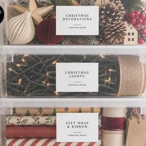 May include: Three clear plastic storage bins stacked vertically, each featuring a white rectangular adhesive label. The top bin contains ornaments, the middle bin holds string lights and burlap ribbon, and the bottom bin stores gift wrap and ribbon.