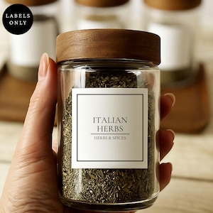 Pode incluir: Um frasco de especiarias de vidro transparente com tampa de madeira escura, cheio de ervas verdes secas. Um rótulo quadrado branco na frente apresenta uma borda preta e o texto "ITALIAN HERBS" acima de "HERBS & SPICES" em fonte preta.