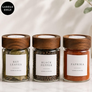 Peut inclure: Trois bocaux à épices en verre transparent avec des couvercles en bois foncé. Chaque bocal porte une étiquette carrée blanche avec le texte noir Bay Leaves, Black Pepper et Paprika, avec la mention Herbs & Spices. Les bocaux sont remplis.