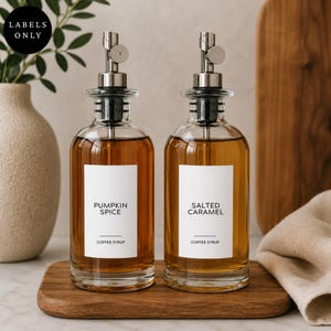 May include: Two clear glass bottles filled with amber liquid, each featuring a silver metal pour spout and a white rectangular label. One label reads PUMPKIN SPICE COFFEE SYRUP, and the other reads SALTED CARAMEL COFFEE SYRUP in black text.