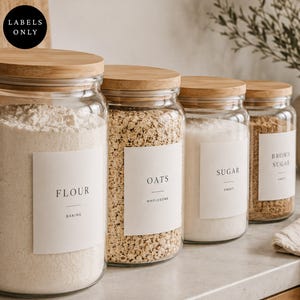 Puede incluir: Cuatro frascos de almacenamiento de vidrio transparente con tapas de madera clara, cada uno con diferentes ingredientes secos. Las etiquetas rectangulares blancas presentan texto negro que dice Flour, Oats, Sugar y Brown Sugar.