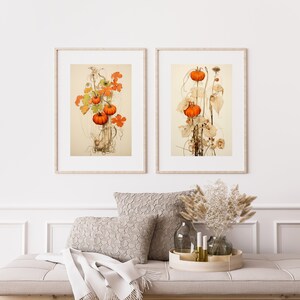 Peut inclure: Deux impressions botaniques encadrées représentant des illustrations détaillées de citrouilles orange avec des feuilles et des vignes vertes. Les impressions sont affichées sur un mur blanc au-dessus d'un canapé brun clair avec des coussins blancs et un vase de fleurs séchées.