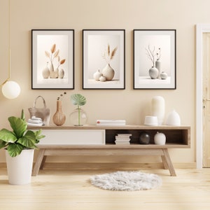 Peut inclure: Une console en bois clair avec trois étagères, un tiroir blanc et un tapis blanc devant. Trois impressions encadrées avec des fonds beiges et des arrangements floraux séchés dans des vases blancs sont accrochés au mur au-dessus de la table.