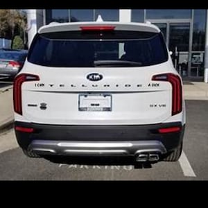 Może przedstawiać: Biały SUV Kia Telluride z czarnymi ozdobami i tylną tablicą rejestracyjną. Zderzak tylny ma dwa rury wydechowe. Tylna szyba ma naklejkę z napisem "Potrafię" i inną naklejkę z napisem "A Dick".