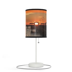 May include: A white table lamp with a cylindrical lampshade featuring a sunset scene over a pier. The lampshade has an orange band at the top and a black band at the bottom. The lamp has a white base and stem, with a pull chain.