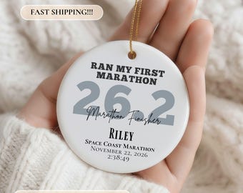 May include: A round white ceramic ornament featuring the black text RAN MY FIRST MARATHON, large grey numbers 26.2, the script text Marathon Finisher, the name RILEY, and the text SPACE COAST MARATHON NOVEMBER 22, 2026 2:38:49 with a gold string.