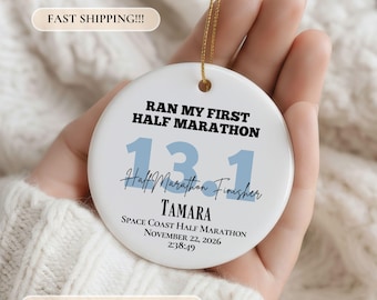 May include: A round white ceramic ornament featuring the black text RAN MY FIRST HALF MARATHON, large light blue numbers 13.1, the script text Half Marathon Finisher, the name TAMARA, and the event details SPACE COAST HALF MARATHON NOVEMBER 22, 2026 2:38:49.