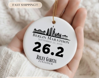 May include: A round white ceramic ornament featuring a black silhouette of the Berlin skyline, the text BERLIN MARATHON, SEPTEMBER 27, 2026, the number 26.2, and the name RILEY GARCIA FINISHER. It hangs from a thin gold string.