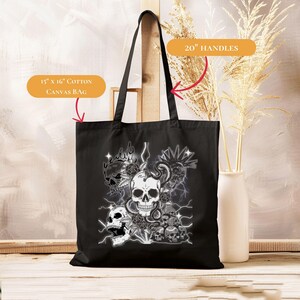 Puede incluir: Bolso tote de lona de algodón negro, 38 cm x 41 cm, con asas de 51 cm. El bolso presenta un gráfico de varias calaveras con rosas, rayos y una serpiente. El diseño es en tonos grises y blancos.