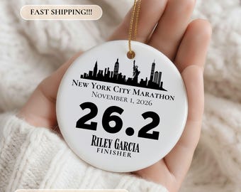 May include: A round white ceramic ornament featuring a black silhouette of the New York City skyline, including the Statue of Liberty. Text reads: New York City Marathon, November 1, 2026, 26.2, Riley Garcia, Finisher. Includes a gold hanging string.