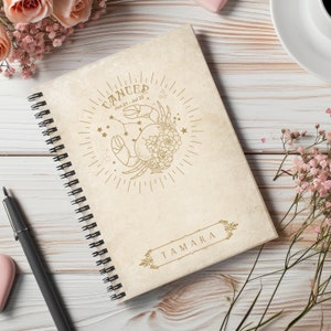 May include: A cream-colored spiral bound notebook with a gold illustration of a crab with flowers and stars. The text "Cancer Jun 21 - Jul 22" is above the illustration. The name "TAMARA" is written in a gold frame at the bottom of the notebook.