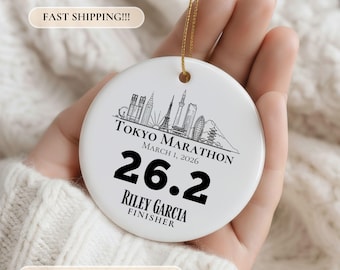 May include: A round white ceramic ornament featuring a black line art illustration of the Tokyo skyline. The text reads TOKYO MARATHON MARCH 1, 2026, 26.2, RILEY GARCIA, and FINISHER. It includes a gold hanging string at the top of the ornament.