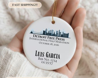 May include: A round white ceramic ornament featuring a blue and black Detroit skyline graphic. Text reads: Detroit Free Press International Half Marathon, October 18, 2026, Luis Garcia, Bib No. 1234, 01:35:57. It hangs from a thin gold string.