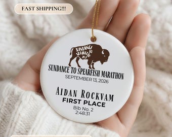 May include: A round white ceramic ornament featuring a brown buffalo silhouette with the text Sundance to Spearfish Marathon 26.2, the date September 13, 2026, the name Aidan Rockvam, the text First Place, and the details Bib No. 2 and 2:48:31.