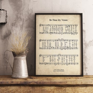 Op de afbeelding: Een ingelijste afdruk van bladmuziek met de titel "Be Thou My Vision". De muziek is gedrukt op verouderd papier met een zwart frame. Een metalen kan met gedroogd gras staat op een houten plank.