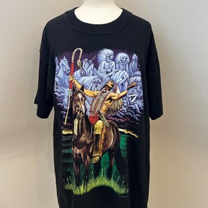 Puede incluir: Una camiseta negra de manga corta con un estampado gráfico de una persona con vestimenta tradicional montando un caballo marrón y sosteniendo un bastón. El fondo muestra otras figuras a caballo bajo un cielo oscuro con hierba verde.