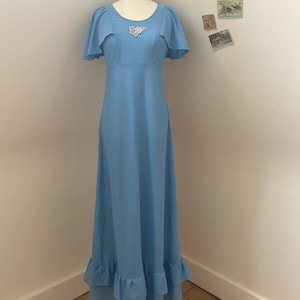 May include: A light blue vintage maxi dress with short flutter sleeves and a ruffled hem. The dress features a decorative white embellishment at the neckline and is displayed on a mannequin. The dress is floor-length and has a fitted bodice.