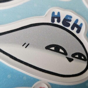 Peut inclure: Un poisson de dessin anim&eacute; blanc et gris avec des contours noirs et une texture scintillante. Le poisson a la bouche ferm&eacute;e et deux yeux. Le poisson dit "HEH" en lettres bleues et noires.
