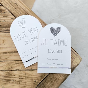 Peut inclure: Deux cartes blanches avec des sommets arqués, chacune avec une illustration de cœur et les phrases "LOVE YOU Je t'aime" et "JE T'AIME Love You" en noir. En dessous de chaque carte se trouve un bracelet avec une barre argentée.