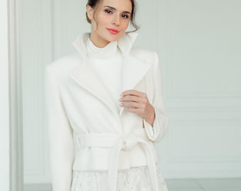 Könnte beinhalten: Eine weiße Jacke mit hohem Kragen und einem Gürtel, der in der Taille gebunden ist. Die Jacke wird über einem weißen Rollkragenpullover und einem weißen Spitzenrock getragen.
