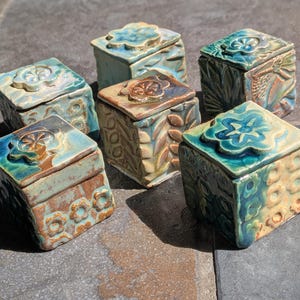 May include: A collection of five square ceramic boxes with lids, each featuring a sculpted flower on top. The boxes are glazed in shades of blue, green, and brown, with embossed floral and leaf patterns on the sides and textured surfaces throughout.