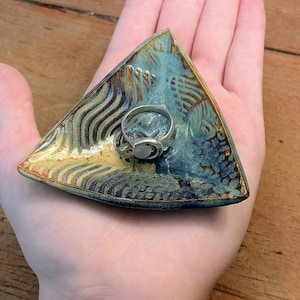 May include: A triangular ceramic trinket dish featuring a blue and gold glaze with textured, wavy patterns. Resting in the center of the dish is a silver ring with a small, round, translucent white stone set into the band.