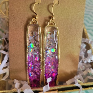 Puede incluir: Un par de pendientes colgantes dorados. Cada pendiente presenta un diseño rectangular de resina transparente lleno de purpurina rosa e iridiscente. Los pendientes tienen un marco y ganchos para las orejas dorados.