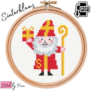 Puede incluir: Un patrón de punto de cruz con Sinterklaas, una figura holandesa similar a Santa Claus, sosteniendo un regalo y un bastón. El patrón está en un aro de bordado de madera circular.