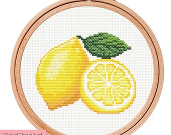 May include: A cross-stitch pattern featuring a whole bright yellow lemon and a sliced lemon half with a green leaf, displayed within a round wooden embroidery hoop. The design is stitched on white fabric with a visible grid texture.