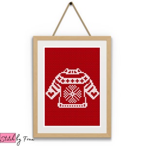 Op de afbeelding: Een ingelijst kruissteekontwerp van een witte kersttrui met een sneeuwvlok op een rode achtergrond.