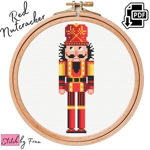 May include: A cross-stitch pattern of a red nutcracker soldier with a gold and black uniform. The pattern is framed in a wooden embroidery hoop. The text "Red Nutcracker" is in the top left corner of the image.