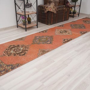 Puede incluir: Una alfombra de pasillo naranja con un patrón repetido de diseños geométricos marrones y beige. La alfombra tiene forma rectangular y está colocada sobre un suelo de madera clara. El borde de la alfombra presenta un patrón azul oscuro y marrón.