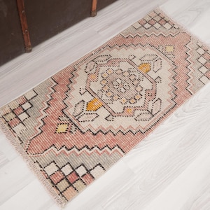 May include: A rectangular, vintage-style rug with a central medallion design. The rug features a color palette of pink, beige, yellow, and black. The edges have a fringed detail. The rug is placed on a light-colored wooden floor.
