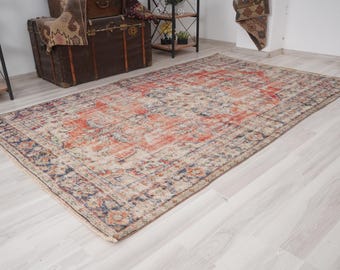 May include: A rectangular, distressed-look rug with a central orange and red design, surrounded by a border of blue and beige. The rug is placed on a light-colored wooden floor, showcasing a vintage aesthetic.