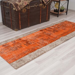 May include: An orange and beige vintage runner rug with a distressed, floral pattern. The rug is rectangular and has a beige border. A dark brown trunk is in the background.