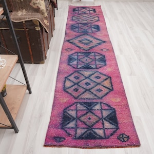 May include: A long, rectangular rug with a vibrant pink background and geometric patterns in navy blue and beige. The rug features repeating diamond and hexagonal shapes, creating a symmetrical design. The rug is placed on a light-coloured wooden floor.