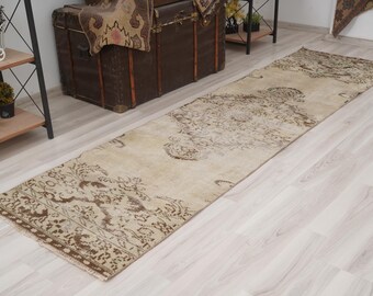 Op de afbeelding: Een rechthoekig, beige loperkleed met een gedetailleerd, bruin bloemenpatroon. Het kleed ligt op een lichtgekleurde houten vloer. De loper is lang en smal, geschikt voor gangen of entrees. Het ontwerp van het kleed is vintage-geïnspireerd.