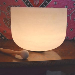 May include: An off-white crystal singing bowl with a wooden mallet. The bowl has a rounded base and a smooth surface. The mallet has a wooden handle and a soft, rounded head. The bowl and mallet are on a dark wooden surface.
