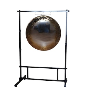 May include: A large, bronze-coloured gong with a textured surface, suspended from a black metal stand. The gong is round and reflects the surrounding environment. The stand has a rectangular base and a horizontal bar.