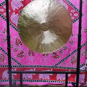 May include: A large, round, gold-coloured gong hanging from a black metal stand. The stand has wheels and is in front of a pink and black patterned fabric.