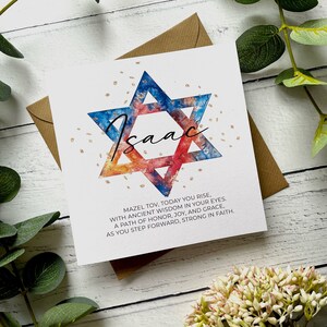 May include: A white card featuring a watercolour Star of David in blue, red, and gold, with the name "Isaac" in cursive. The card includes the text "Mazel Tov, Today You Rise..." and is set on a brown envelope.