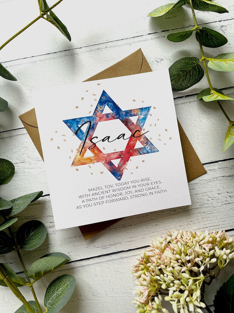 May include: A white card featuring a watercolour Star of David in blue, red, and gold, with the name "Isaac" in cursive. The card includes the text "Mazel Tov, Today You Rise..." and is set on a brown envelope.