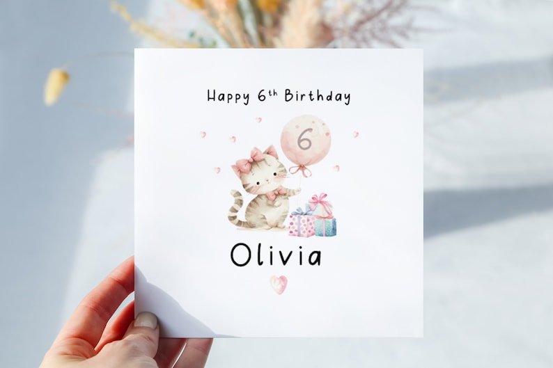 May include: A white birthday card with a watercolor illustration of a kitten holding a balloon with the number 6. The card reads "Happy 6th Birthday Olivia" with pink accents and gift boxes.