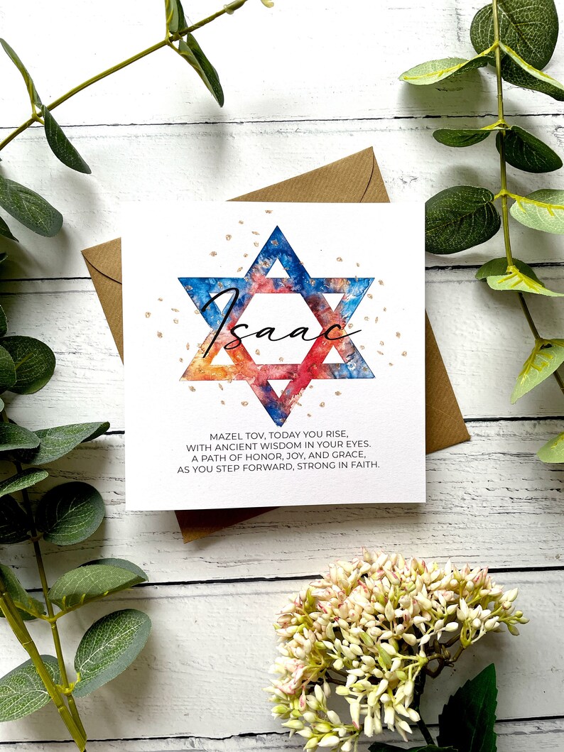 May include: A greeting card featuring a watercolour Star of David in blue, orange, and gold. The name "Isaac" is written in cursive. The card includes the text: "Mazel Tov, today you rise..." and is placed on a brown envelope.