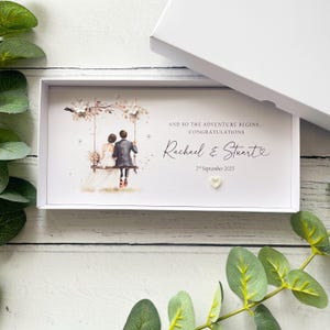 May include: A rectangular white gift box containing a personalized card with an illustration of a couple on a swing. The text reads: AND SO THE ADVENTURE BEGINS... CONGRATULATIONS, Rachael & Stuart, 2nd September 2025. Includes small white floral accents.