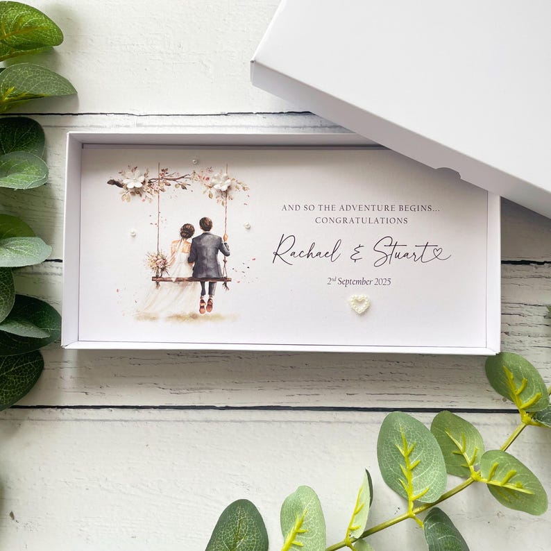 May include: A rectangular white gift box containing a personalized card with an illustration of a couple on a swing. The text reads: AND SO THE ADVENTURE BEGINS... CONGRATULATIONS, Rachael & Stuart, 2nd September 2025. Includes small white floral accents.