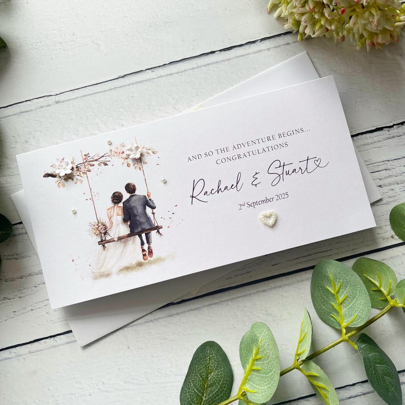 May include: A white rectangular wedding card featuring a watercolor illustration of a couple on a swing, with the text AND SO THE ADVENTURE BEGINS... CONGRATULATIONS, the names Rachael & Stuart, the date 2nd September 2025, and a small heart.