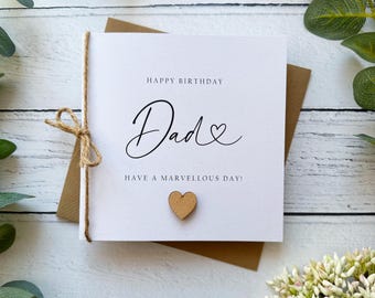 May include: A white greeting card with the words "Happy Birthday Dad" in elegant script, a small wooden heart, and a twine bow. The card is on a brown envelope, with the words "Have a Marvellous Day!"